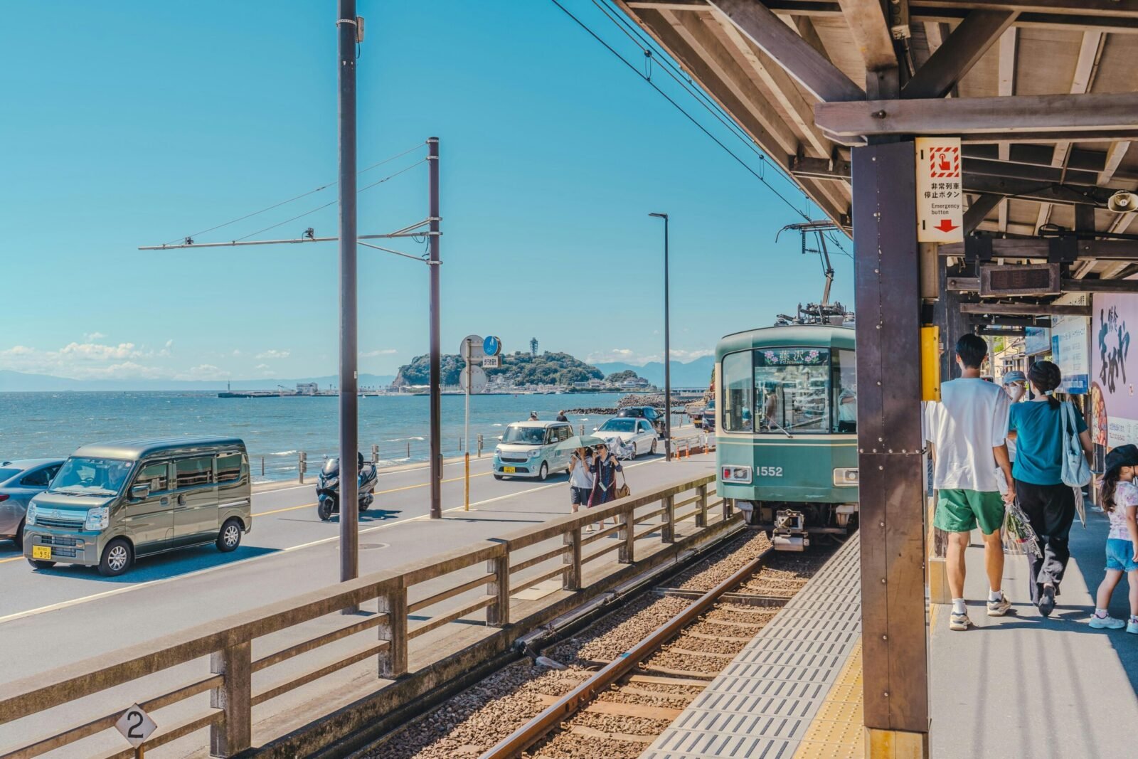 A vintage green and cream Enoden train pulling up to a sunny outdoor station platform next to a coastal road and the blue sea in Kamakura, Japan.