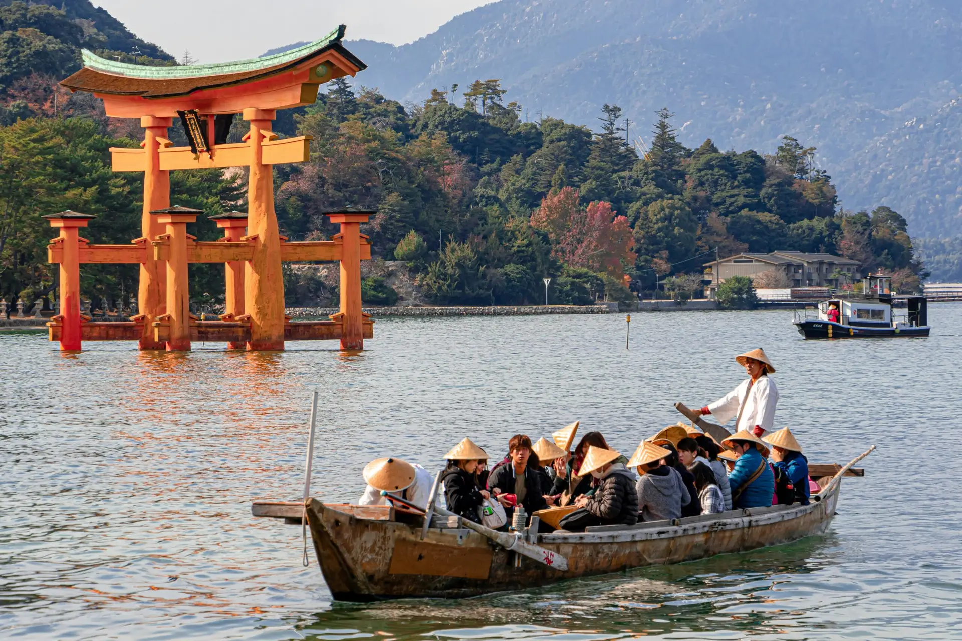 A traditional wooden boat filled with tourists wearing conical straw hats floating in the water near the giant orange Itsukushima Shrine Torii gate in Japan.