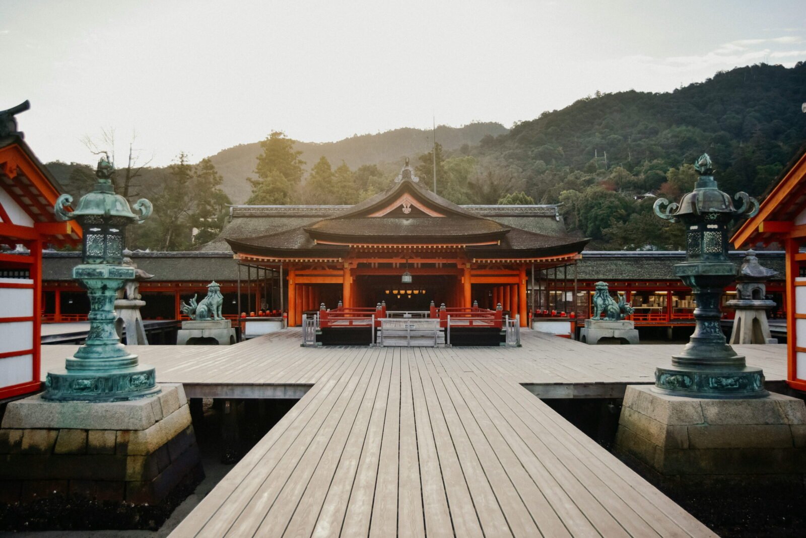 Symmetrical view of the open wooden boardwalk leading toward the red and orange main hall of Itsukushima Shrine in Miyajima, Japan, flanked by large green bronze lanterns.