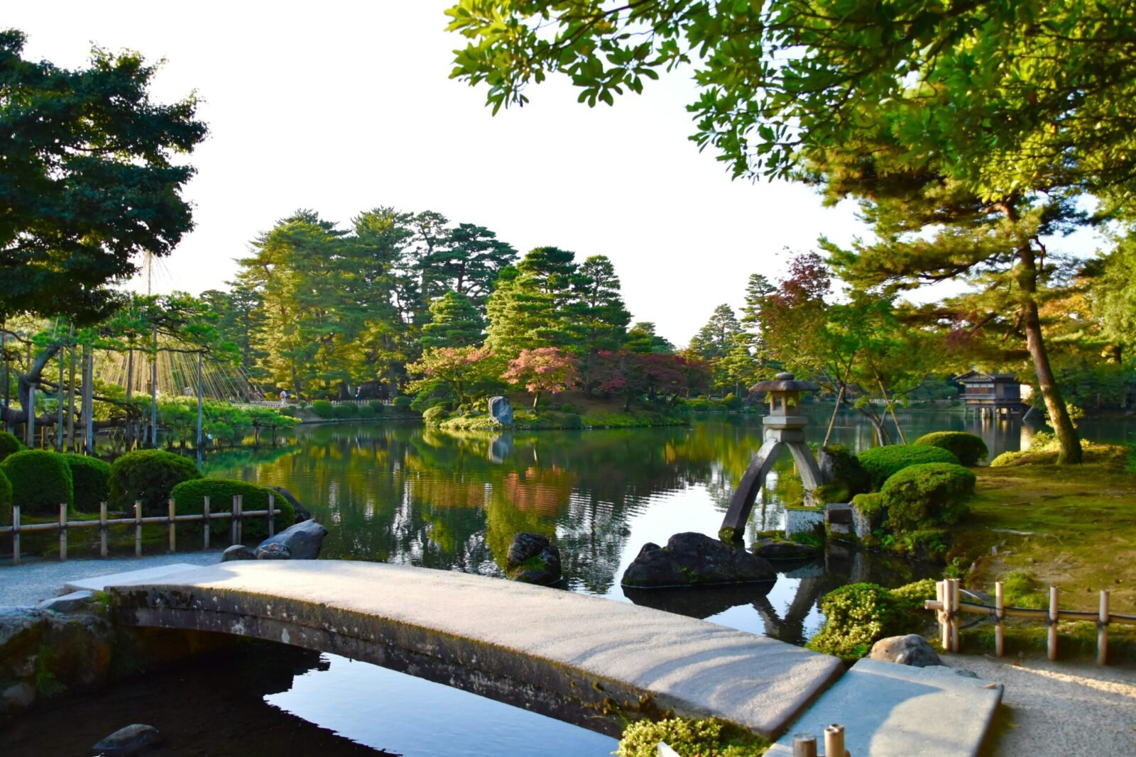 View of a curved stone footbridge stretching over the calm waters of Kasumigaike Pond at Kenroku-en Garden in Kanazawa, Japan, with sculpted pine trees and a two-legged stone lantern.