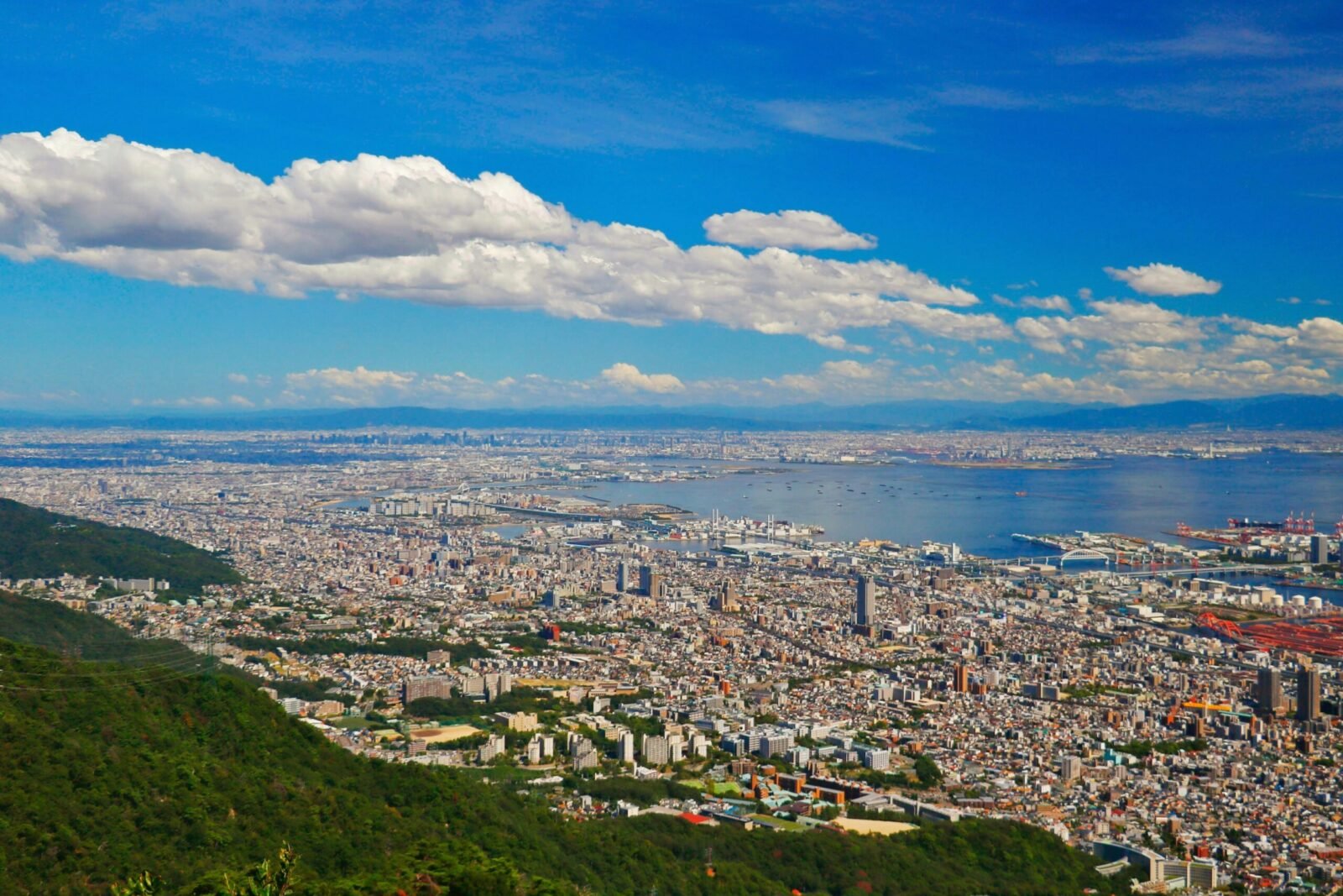 High-angle panoramic view of Kobe city's dense urban layout sprawling down toward the blue waters of Osaka Bay under a bright blue sky.