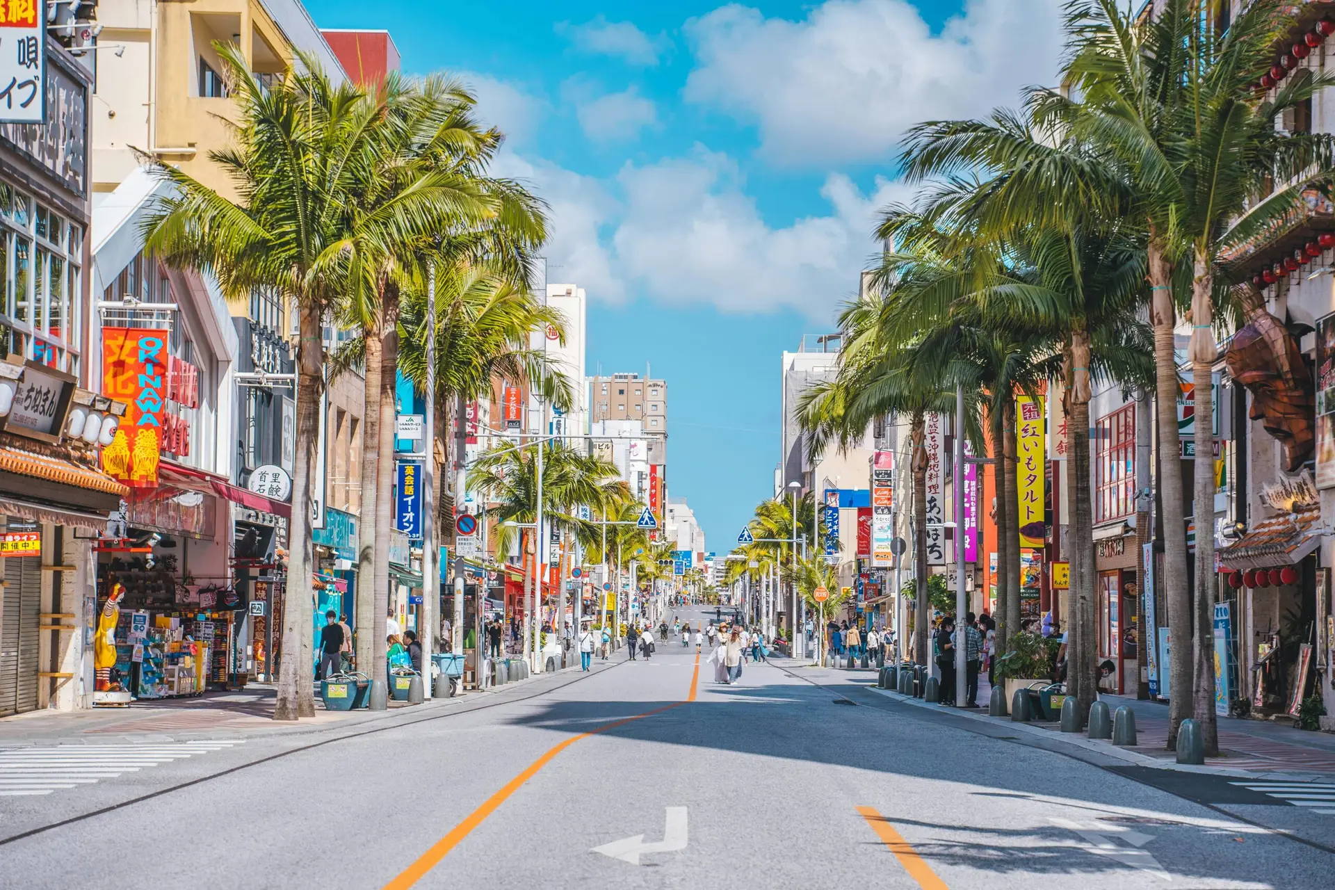 View looking down a bright, sunlit city street lined with tall palm trees, shops, and colorful signs under a blue sky in Naha, Okinawa.